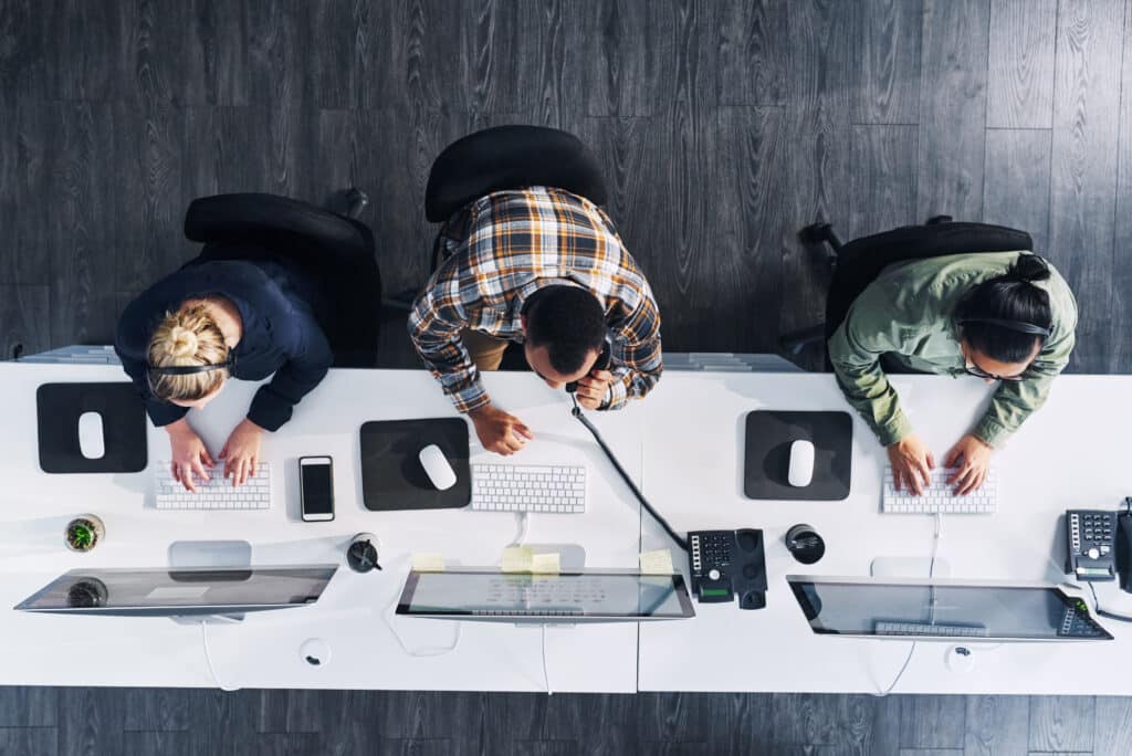 An arial view of three customer service agents. They sit in front of their computers typing. One of the customer service agents is taking a call.