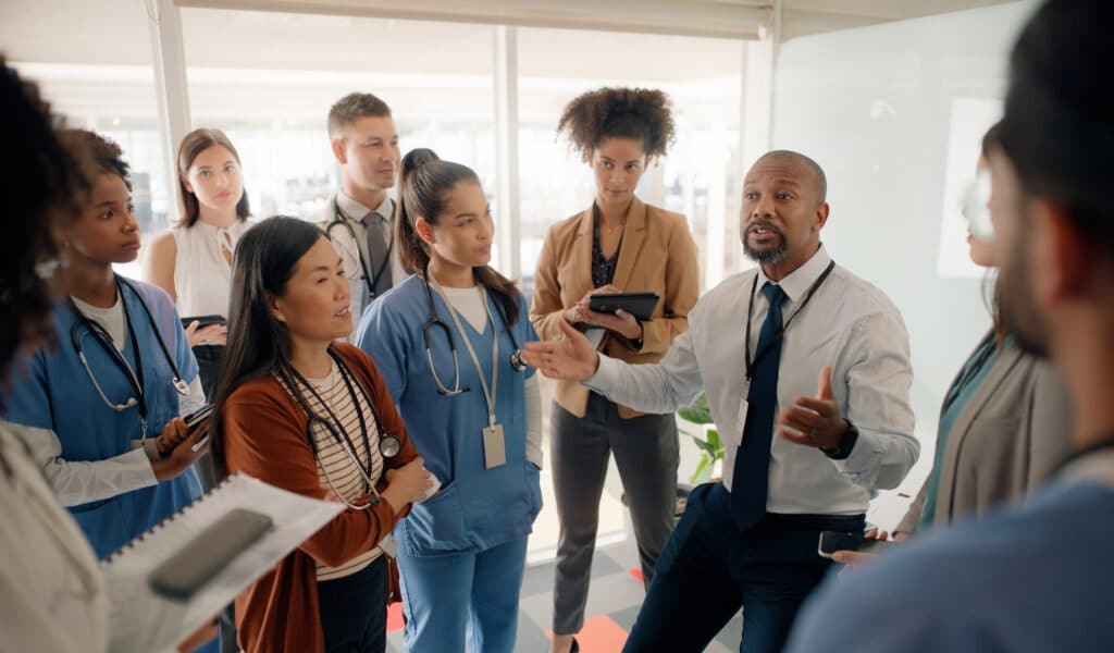 A group of healthcare professionals, some in scrubs and wearing stethoscopes and name badges, taking notes gathered around a man who is speaking.