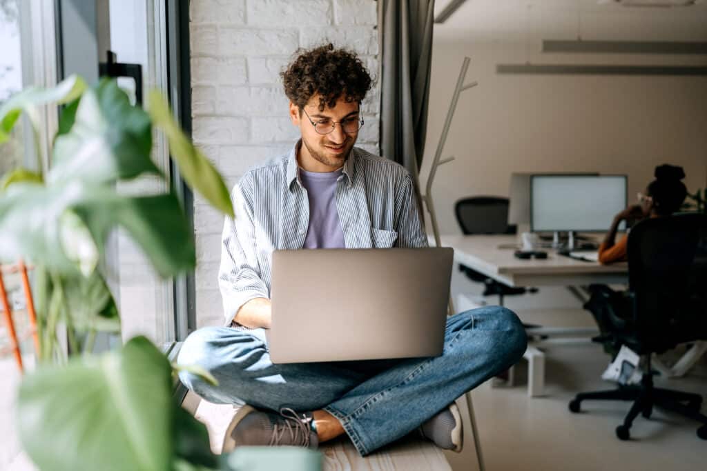 A casually dressed man sits in a window and works on a computer.